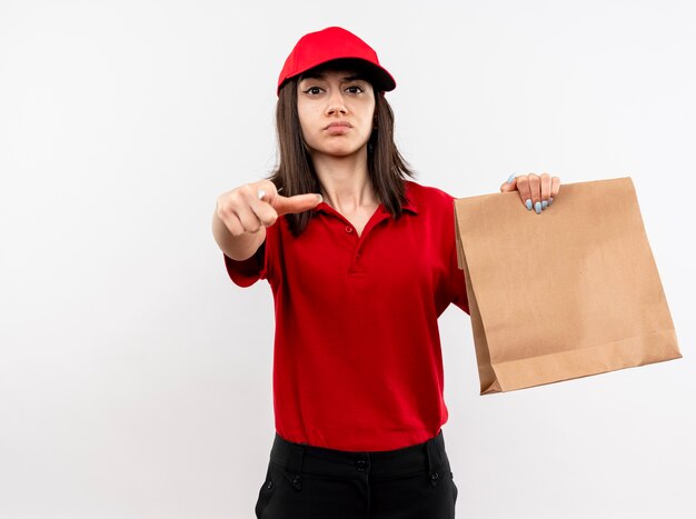 Jeune livreuse portant l'uniforme rouge et cap tenant le paquet de papier pointant avec l'index à la caméra à la face sérieuse debout sur fond blanc