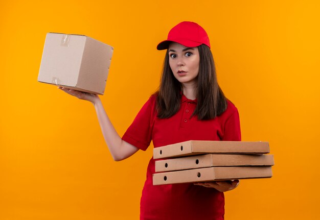 Jeune livreuse surprise portant un t-shirt rouge en bonnet rouge tenant la boîte et la boîte à pizza sur fond jaune isolé