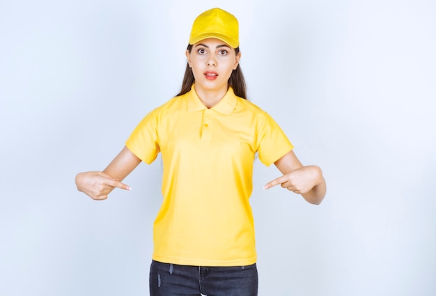 Photo gratuite jeune livreuse en t-shirt jaune et casquette pointant sur elle-même sur blanc.