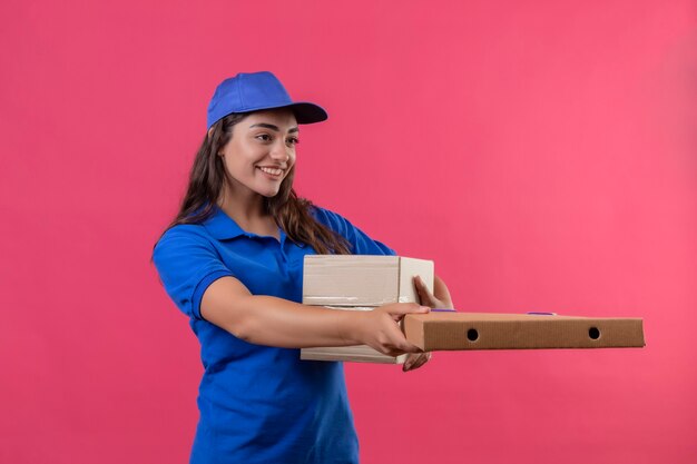 Jeune livreuse en uniforme bleu et cap donnant des boîtes en carton à un client souriant sympathique debout sur fond rose