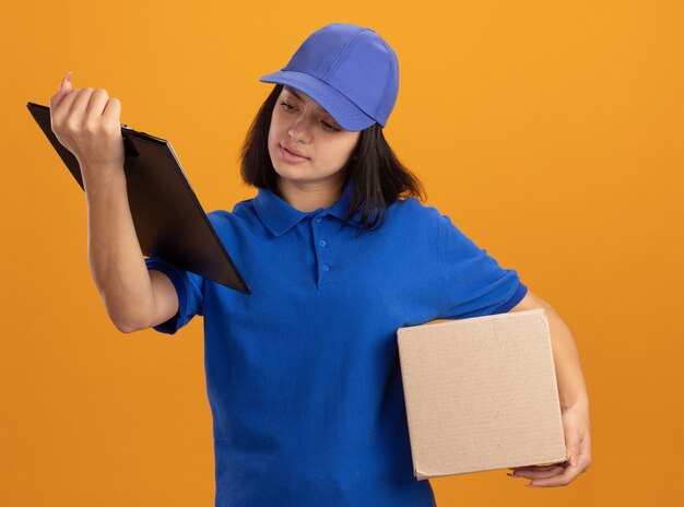 Jeune livreuse en uniforme bleu et cap holding boîte en carton et presse-papiers avec des pages vierges à la recherche de pages avec un visage sérieux debout sur un mur orange