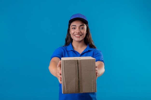 Jeune livreuse en uniforme bleu et cap holding box package le donnant à un client debout sur fond bleu