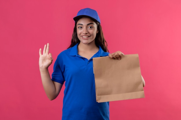 Jeune Livreuse En Uniforme Bleu Et Cap Holding Paper Package Looking At Camera Smiling Friendly Faisant Signe Ok Debout Sur Fond Rose