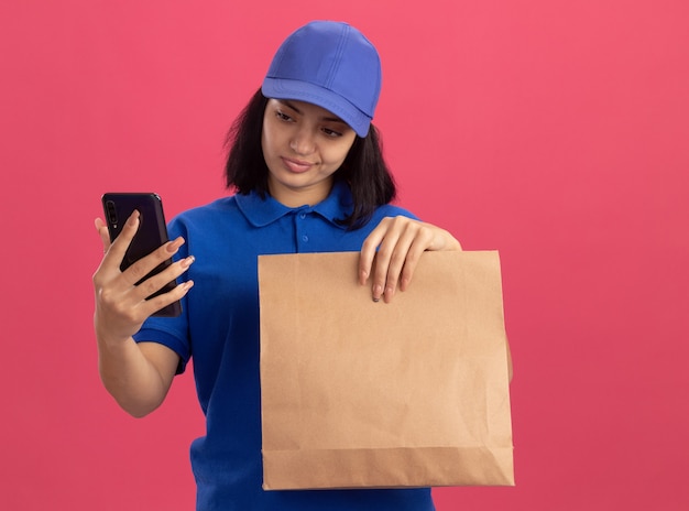 Jeune livreuse en uniforme bleu et cap holding paper package regardant son smartphone avec une expression sceptique debout sur un mur rose