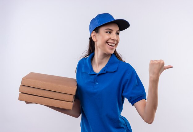 Jeune livreuse en uniforme bleu et cap holding pile de boîtes de pizza pointant vers l'arrière avec le pouce souriant joyeusement