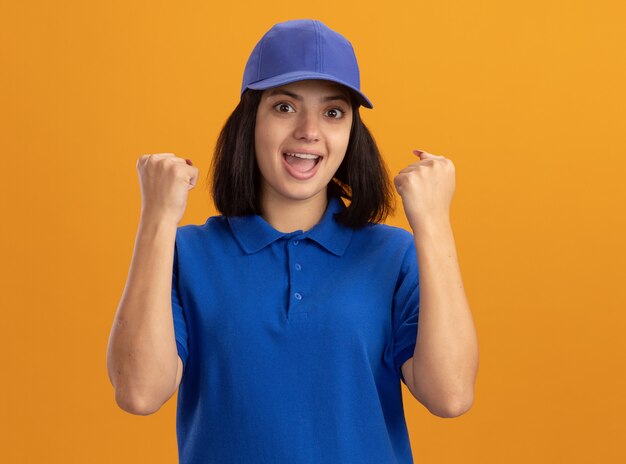 Jeune livreuse en uniforme bleu et cap serrant les poings hapy et excité debout sur le mur orange