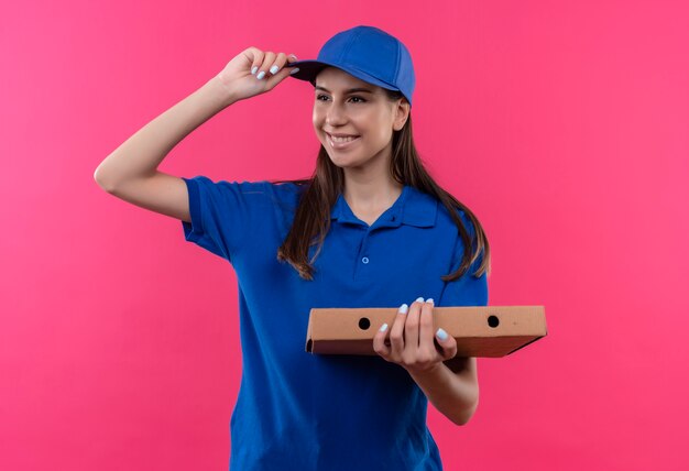 Jeune livreuse en uniforme bleu et cap tenant la boîte à pizza à côté souriant joyeusement