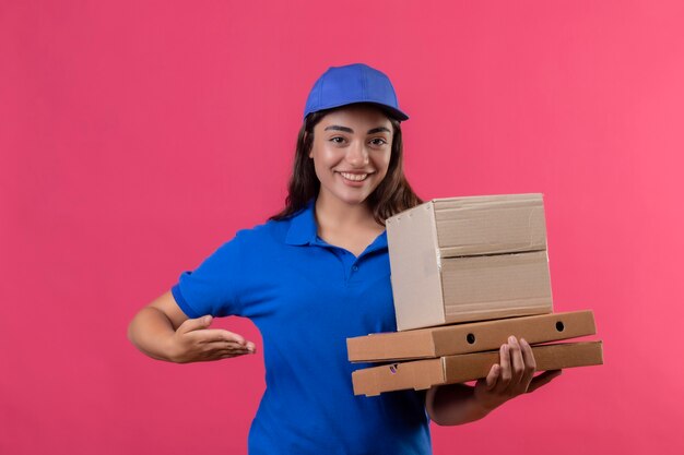 Jeune livreuse en uniforme bleu et cap tenant des boîtes à pizza et boîte de présentation avec bras de main souriant joyeusement heureux et positif debout sur fond rose