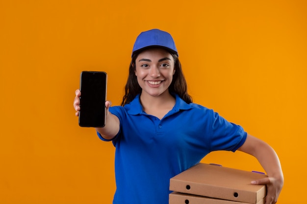 Jeune livreuse en uniforme bleu et cap tenant des boîtes à pizza montrant smartphone à caméra souriant sympathique debout sur fond jaune