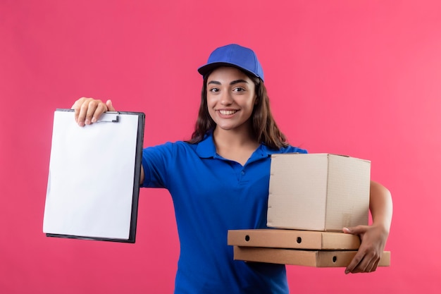 Jeune livreuse en uniforme bleu et cap tenant des boîtes à pizza et un paquet de boîte montrant le presse-papiers en regardant la caméra souriant sympathique debout sur fond rose