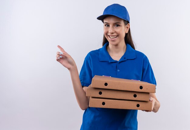 Jeune livreuse en uniforme bleu et cap tenant des boîtes de pizza pointant avec le doigt sur le côté souriant largement avec un visage heureux