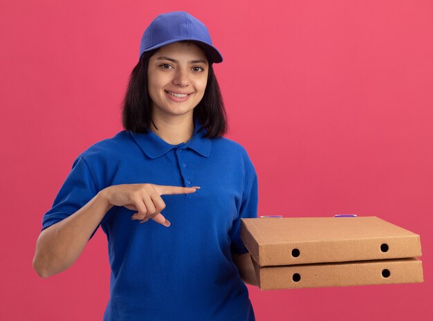 Jeune livreuse en uniforme bleu et cap tenant des boîtes de pizza pointant avec l'index sur le côté souriant debout sur le mur rose