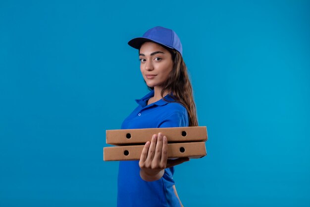 Jeune livreuse en uniforme bleu et cap tenant des boîtes à pizza regardant la caméra souriant confiant heureux et positif debout sur fond bleu