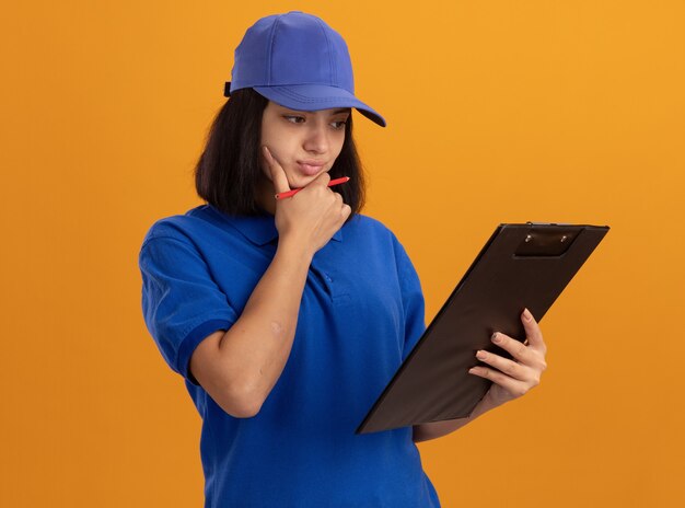 Jeune livreuse en uniforme bleu et cap tenant le presse-papiers en le regardant avec une expression pensive debout sur un mur orange