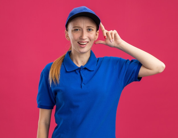 Jeune livreuse en uniforme bleu et casquette confondue avec la main sur la tête debout sur le mur rose