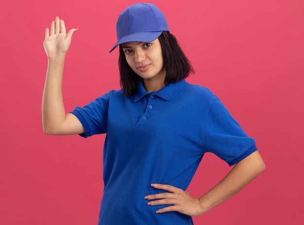 Jeune livreuse en uniforme bleu et casquette avec expression confiante en agitant avec la main debout sur le mur rose