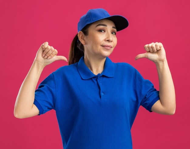 Jeune livreuse en uniforme bleu et casquette avec une expression confiante se montrant avec les pouces debout sur le mur rose