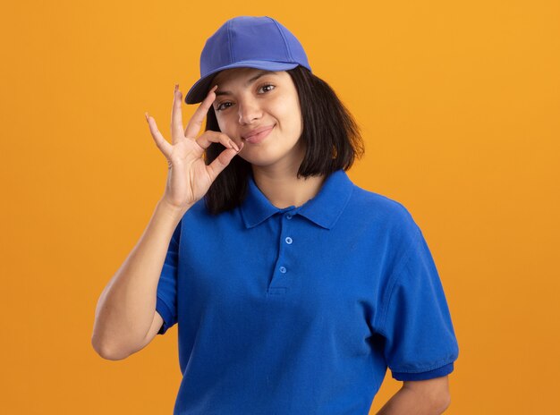 Jeune livreuse en uniforme bleu et casquette faisant un geste de silence comme la fermeture de la bouche avec une fermeture à glissière debout sur un mur orange