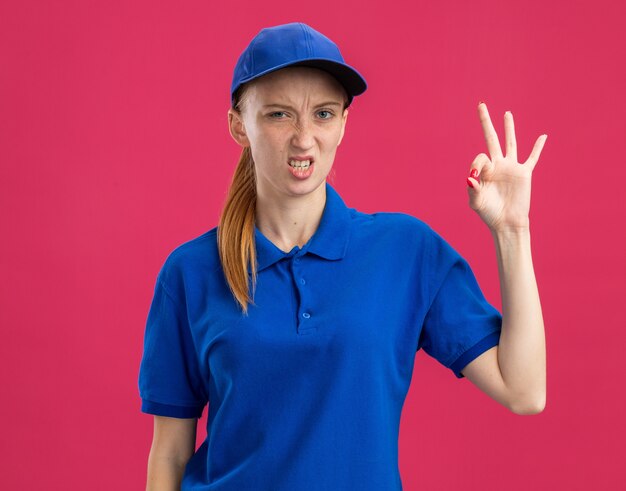 Jeune livreuse en uniforme bleu et casquette mécontente montrant un signe ok debout sur un mur rose
