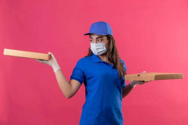 Jeune livreuse en uniforme bleu et casquette portant un masque de protection du visage et des gants tenant des boîtes de pizza à côté avec un visage sérieux debout sur fond rose