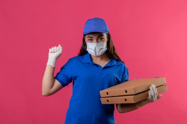 Jeune livreuse en uniforme bleu et casquette portant un masque de protection du visage et des gants tenant des boîtes de pizza levant le poing se réjouissant de son succès et de sa victoire heureux et positif souriant debout sur