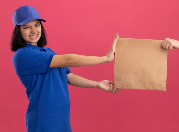 Jeune livreuse en uniforme bleu et casquette refusant de prendre un paquet de papier d'être insatisfait debout sur un mur rose