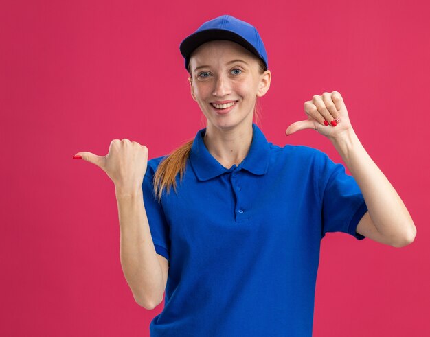 Jeune livreuse en uniforme bleu et casquette souriante confiante pointant les pouces sur le côté debout sur le mur rose