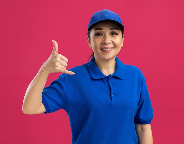 Jeune livreuse en uniforme bleu et casquette avec le sourire sur le visage me faisant appeler le geste debout sur le mur rose