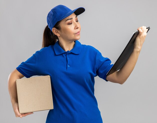 Jeune livreuse en uniforme bleu et casquette tenant une boîte en carton et un presse-papiers le regardant avec un visage sérieux debout sur un mur blanc