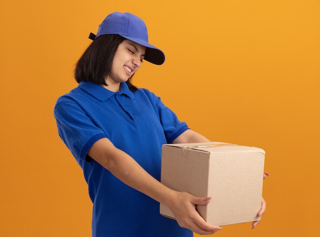 Jeune livreuse en uniforme bleu et casquette tenant une boîte en carton à la regarder avec une expression dégoûtée debout sur un mur orange