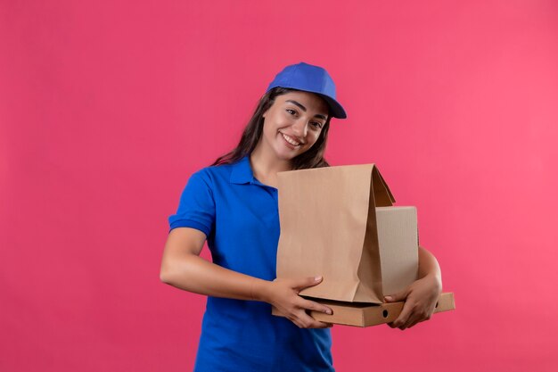 Jeune livreuse en uniforme bleu et casquette tenant des boîtes en carton souriant joyeusement debout sur fond rose