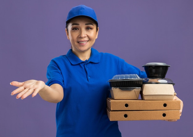 Jeune livreuse en uniforme bleu et casquette tenant des boîtes à pizza et des colis alimentaires avec le bras tendu souriant joyeusement debout sur un mur violet