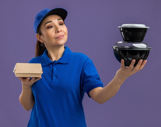 Jeune livreuse en uniforme bleu et casquette tenant des colis alimentaires les regardant avec un sourire sur le visage debout sur un mur violet