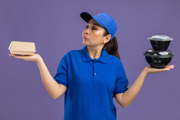 Photo gratuite jeune livreuse en uniforme bleu et casquette tenant des colis alimentaires semblant confuse n'ayant pas de réponse debout sur un mur violet