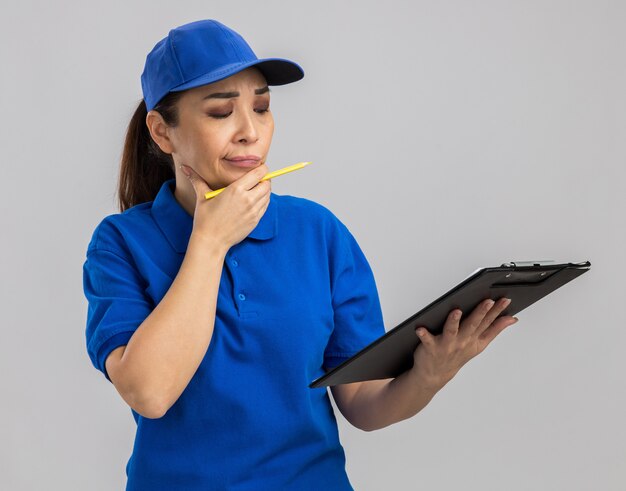 Jeune livreuse en uniforme bleu et casquette tenant un presse-papiers et un stylo regardant avec une expression pensive pensant debout sur un mur blanc