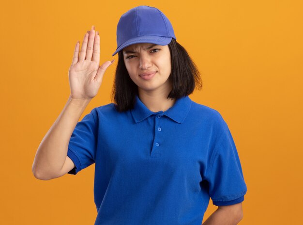 Jeune livreuse en uniforme bleu et casquette avec visage sérieux montrant la main ouverte debout sur le mur orange