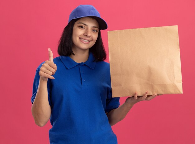 Jeune livreuse en uniforme bleu et chapeau montrant le paquet de papier souriant confiant montrant les pouces vers le haut debout sur le mur rose