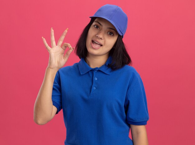 Jeune livreuse en uniforme bleu et chapeau souriant joyeusement montrant signe ok debout sur le mur rose