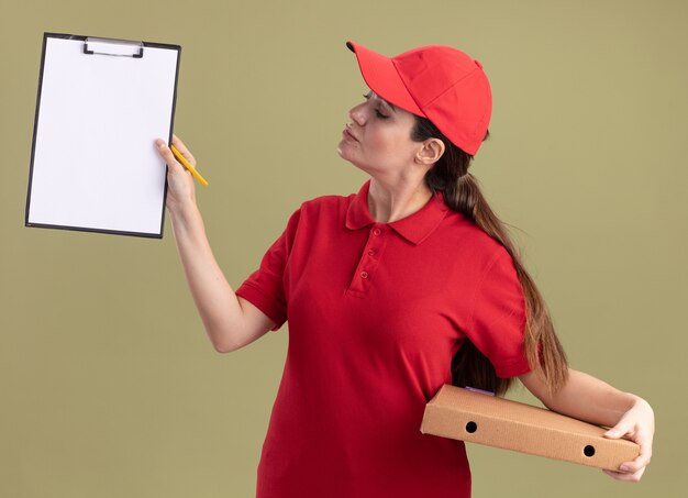 Jeune livreuse en uniforme et casquette tenant un paquet de pizza montrant le presse-papiers avec un crayon à la main en regardant le presse-papiers isolé sur un mur vert olive