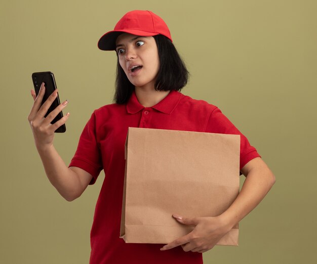 Jeune livreuse en uniforme rouge et cap holding paper package regardant son smartphone avec une expression agacée debout sur un mur léger