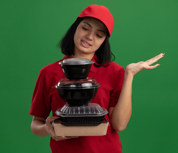 Jeune livreuse en uniforme rouge et cap holding pile de colis alimentaires à la recherche de confus avec bras levé debout sur mur vert