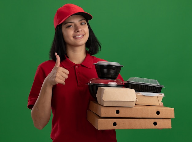 Jeune livreuse en uniforme rouge et cap tenant des boîtes de pizza et des emballages alimentaires avec sourire sur le visage montrant les pouces vers le haut debout sur le mur vert