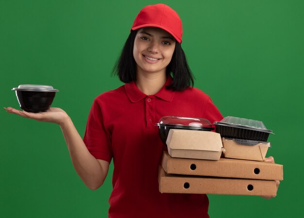 Jeune livreuse en uniforme rouge et cap tenant des boîtes de pizza et des emballages alimentaires wioth sourire sur le visage debout sur le mur vert