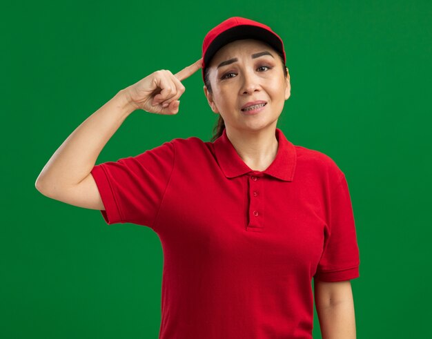 Jeune livreuse en uniforme rouge et casquette confuse et mécontente pointant l'index vers la tête debout sur un mur vert