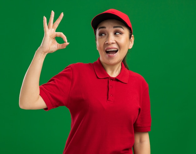 Jeune livreuse en uniforme rouge et casquette regardant de côté heureuse et joyeuse faisant signe ok debout sur un mur vert