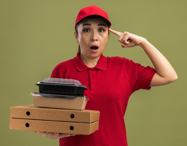 Jeune livreuse en uniforme rouge et casquette tenant des boîtes à pizza et des colis alimentaires pointant avec l'index vers le temple, l'air surpris debout sur un mur vert