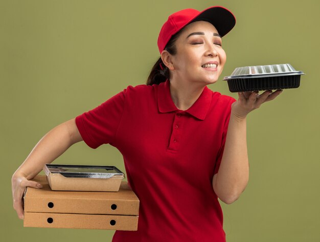 Jeune Livreuse En Uniforme Rouge Et Casquette Tenant Des Boîtes à Pizza Et Des Emballages Alimentaires Inhalant Un Arôme Agréable