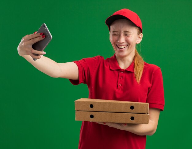 Jeune livreuse en uniforme rouge et casquette tenant des boîtes à pizza faisant du selfie à l'aide d'un smartphone souriant joyeusement debout sur un mur vert