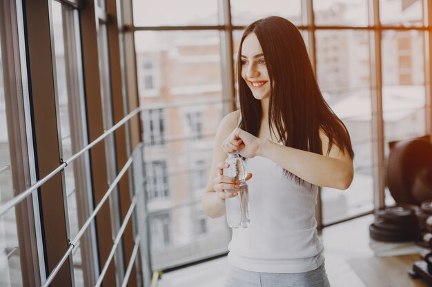 jeune et maigre fille dans une chemise blanche et des leggings gris, debout dans une salle de sport avec une bouteille