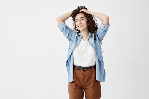 Jeune mannequin brune joyeuse portant un pantalon marron et une chemise en jean, avec une expression rêveuse et joyeuse, jouant avec de longs cheveux ondulés. Jeunesse et beauté